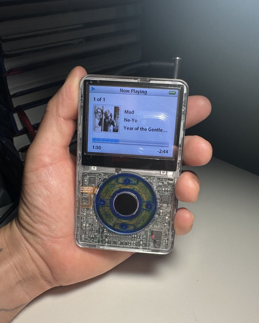 A clear-shell iPod Classic held in a hand, the circuit board visible behind a blue click wheel and a song on screen.
