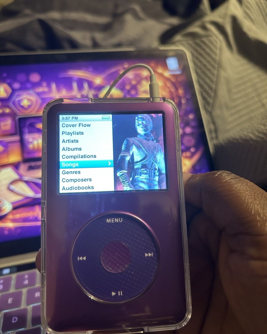 A purple-tinted clear iPod Classic in a hand, lit by a laptop screen, the music menu open.